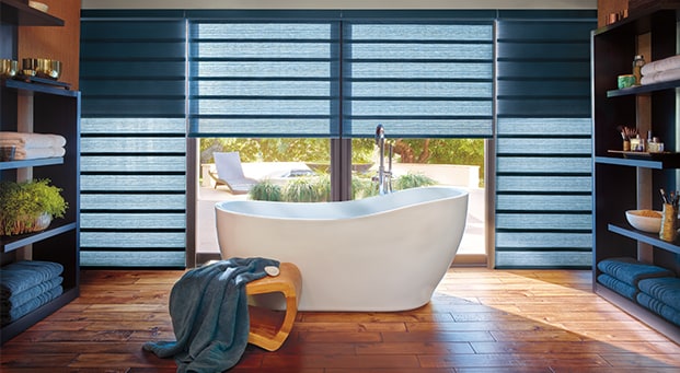 A modern bathroom with a white tub, blue blinds, and wooden floor.